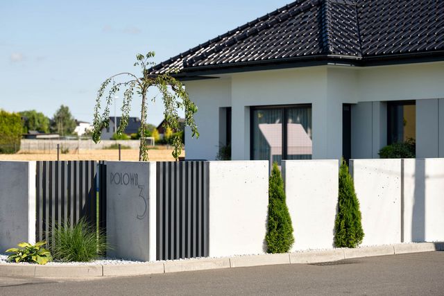 Betonowe ogrodzenie z drewnianymi elementami. Na ogrodzeniu sprefabrykowany jest numer domu oraz nazwa ulicy.