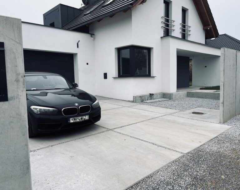 Samochód na betonowym podjeździe do garażu. W tle biały dom ogrodzony betonowym ogrodzeniem.