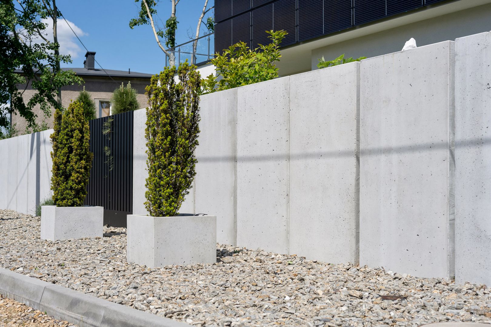 Ogrodzenie wykonane z płyt betonowych. Podsypane kamieniem, w tle dwie duże betonowe donice z dwoma drzewkami.