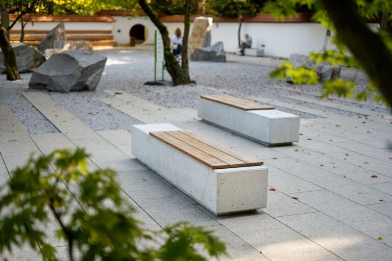 Betonowa ławka z drewnianym siedziskiem w parku, płyty betonowe w tle obsypane liśćmi.